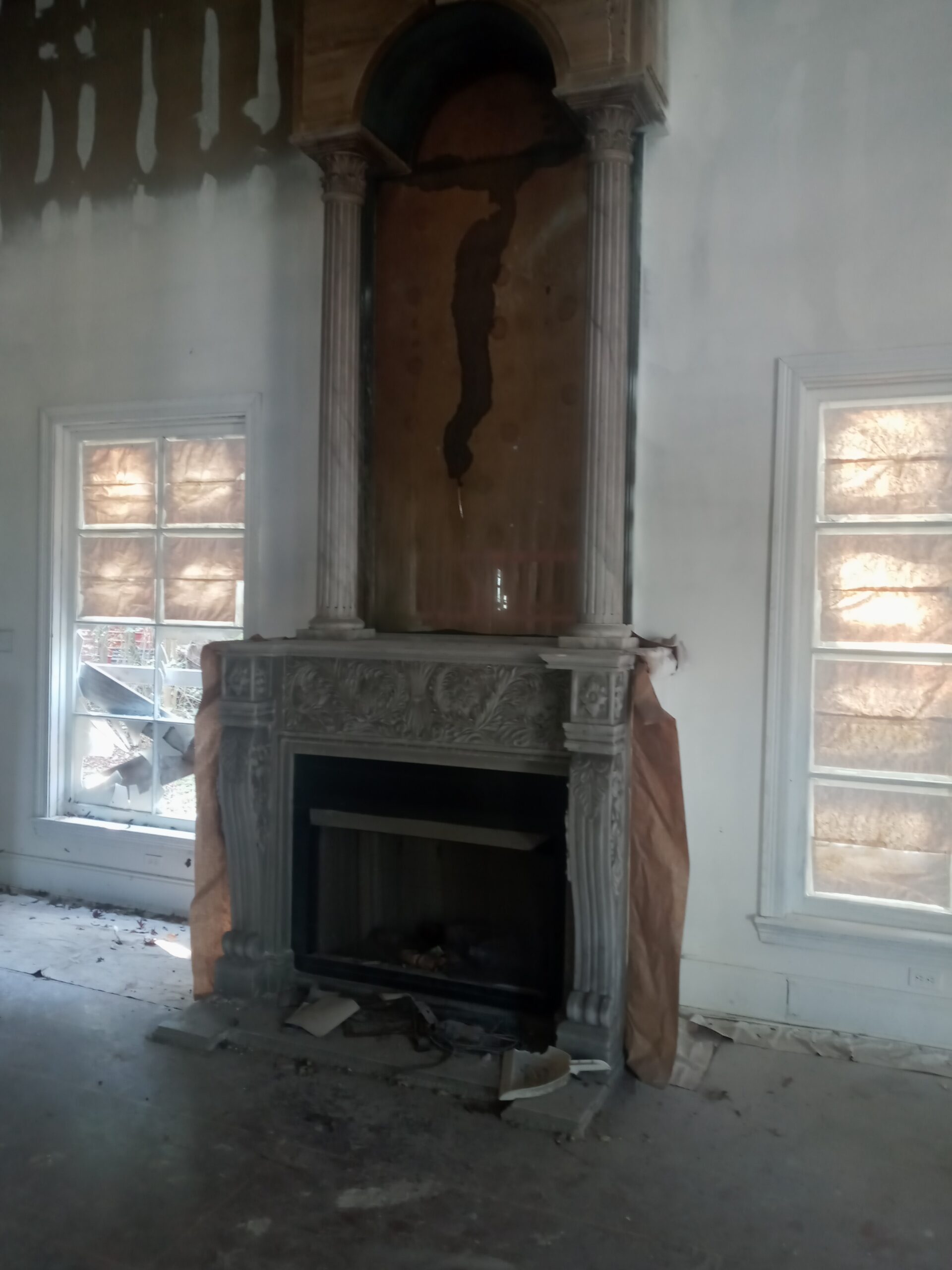 An ornate, empty fireplace surrounded by walls with peeling paint and sunlight filtering through dusty windows.