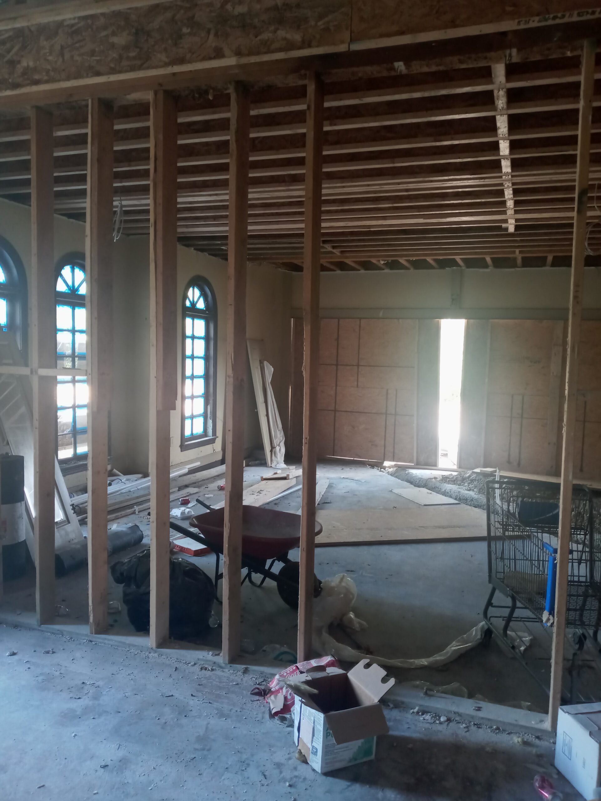 A partially constructed interior space with exposed framing, scattered construction materials, and windows allowing natural light to enter.