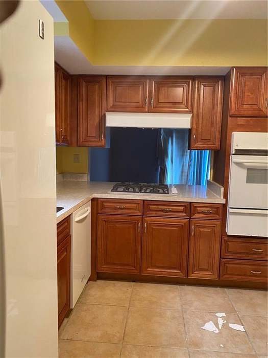 A tidy kitchen featuring wooden cabinets, a white dishwasher, and a gas stove, with light-colored tiles and natural light coming through a window.