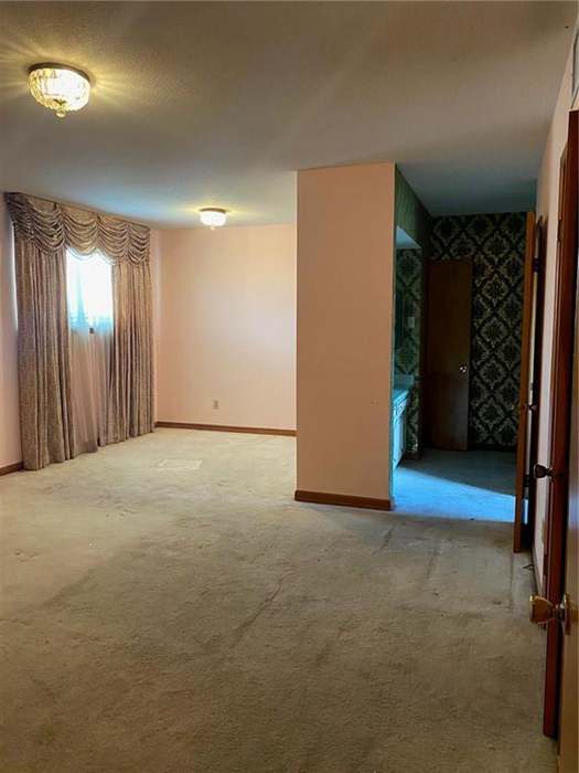 A spacious room with light pink walls, carpeted floor, and curtains, featuring a doorway leading to another area with patterned wallpaper.