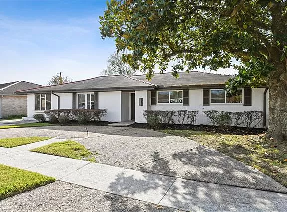 A single-story house with white exterior and a well-maintained yard, featuring a large tree and a driveway leading to the entrance.