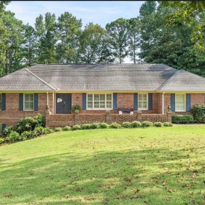 A brick house with a sloping lawn, surrounded by trees, featuring a front porch and multiple windows.