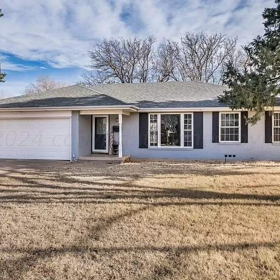 A single-story house with a light grey exterior, a front porch, and several large windows, surrounded by a grassy yard and trees.
