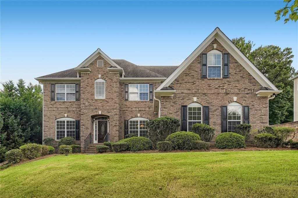 A large, two-story brick house with multiple windows and a lush green lawn in front, framed by trees and a clear blue sky.