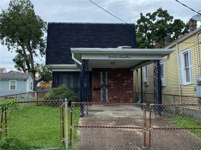 A two-story house with a dark roof, brick facade, and fenced yard, situated in a suburban area with nearby houses.