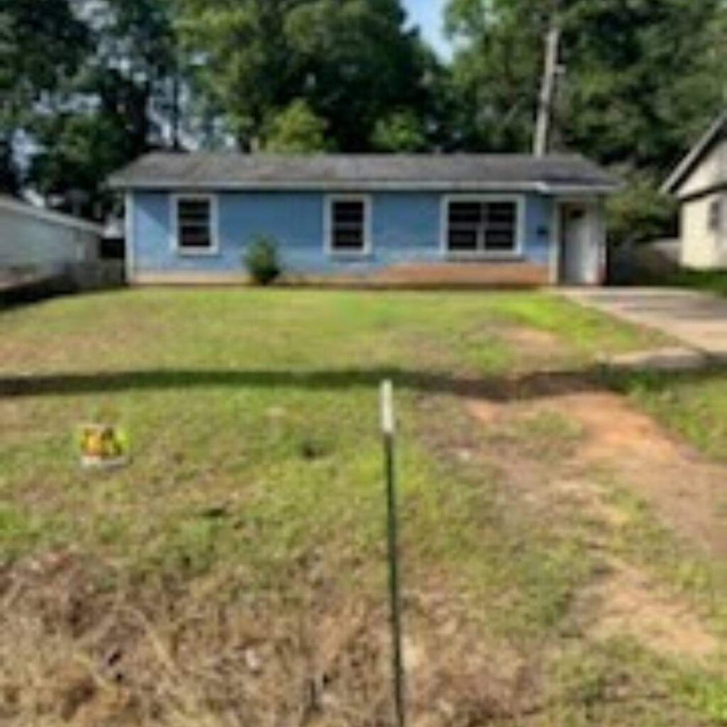 A single-story blue house with a lawn, framed by trees, and a clear blue sky. A driveway is visible alongside the front yard.